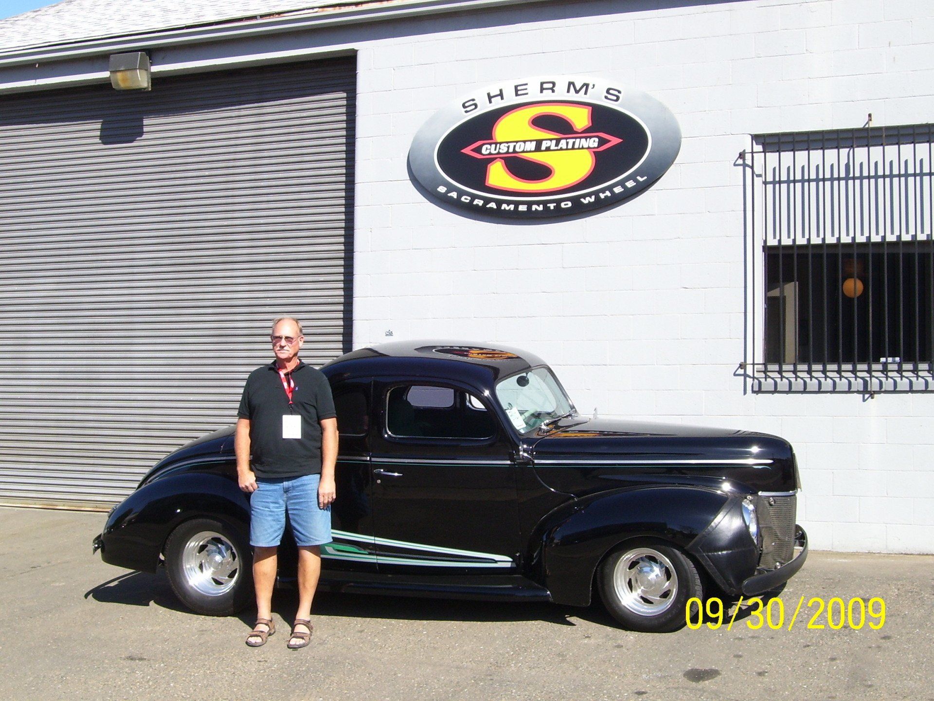 A man standing next to a black car in front of a sign that says charms