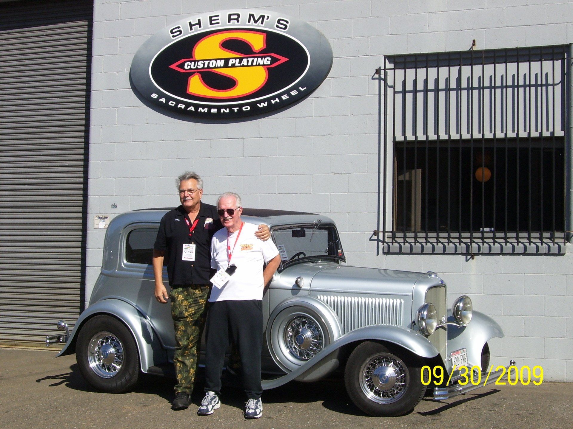 Two men standing next to a silver car in front of a sign that says sherms