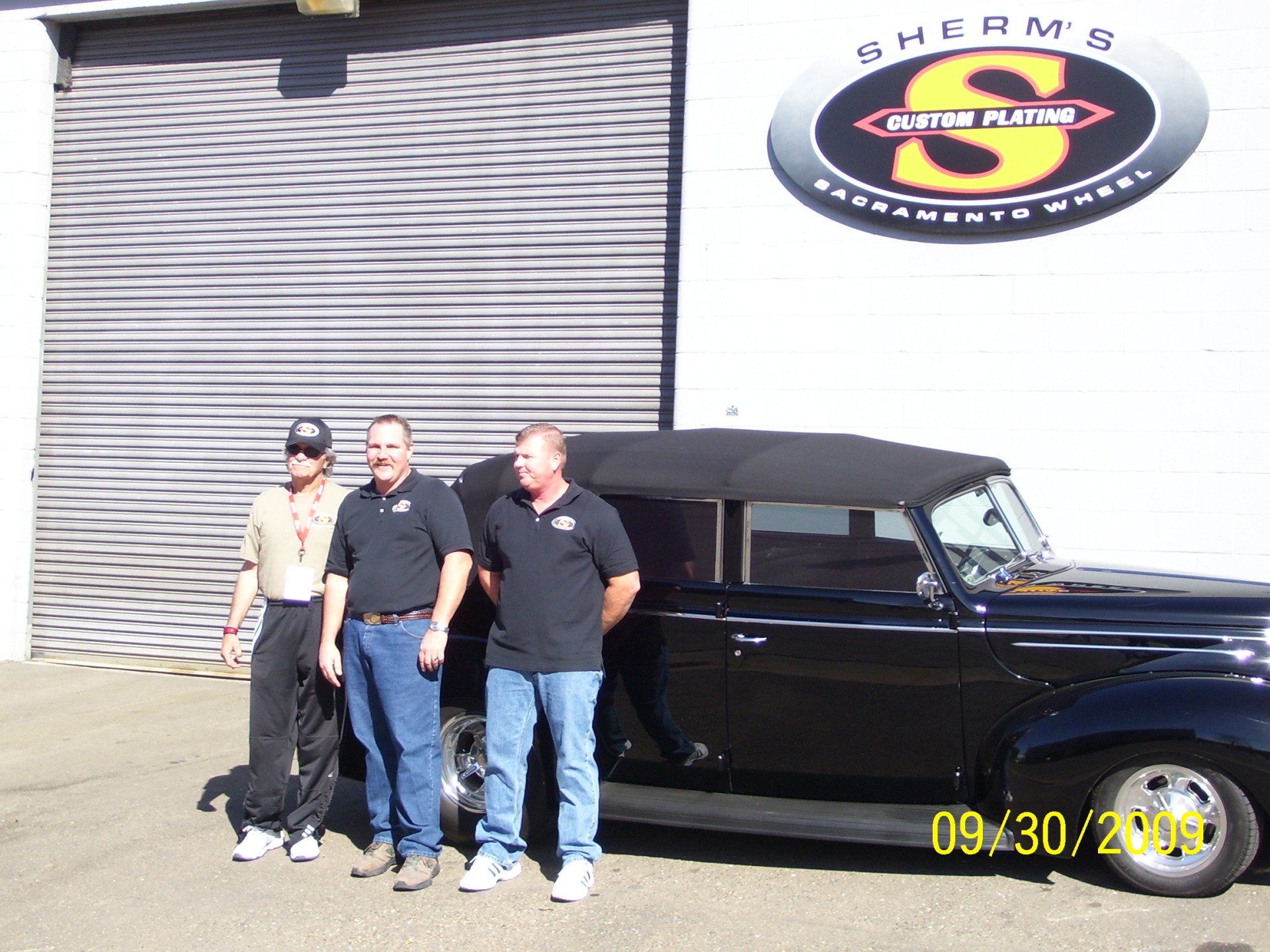 Three men standing in front of a black car that says sherms