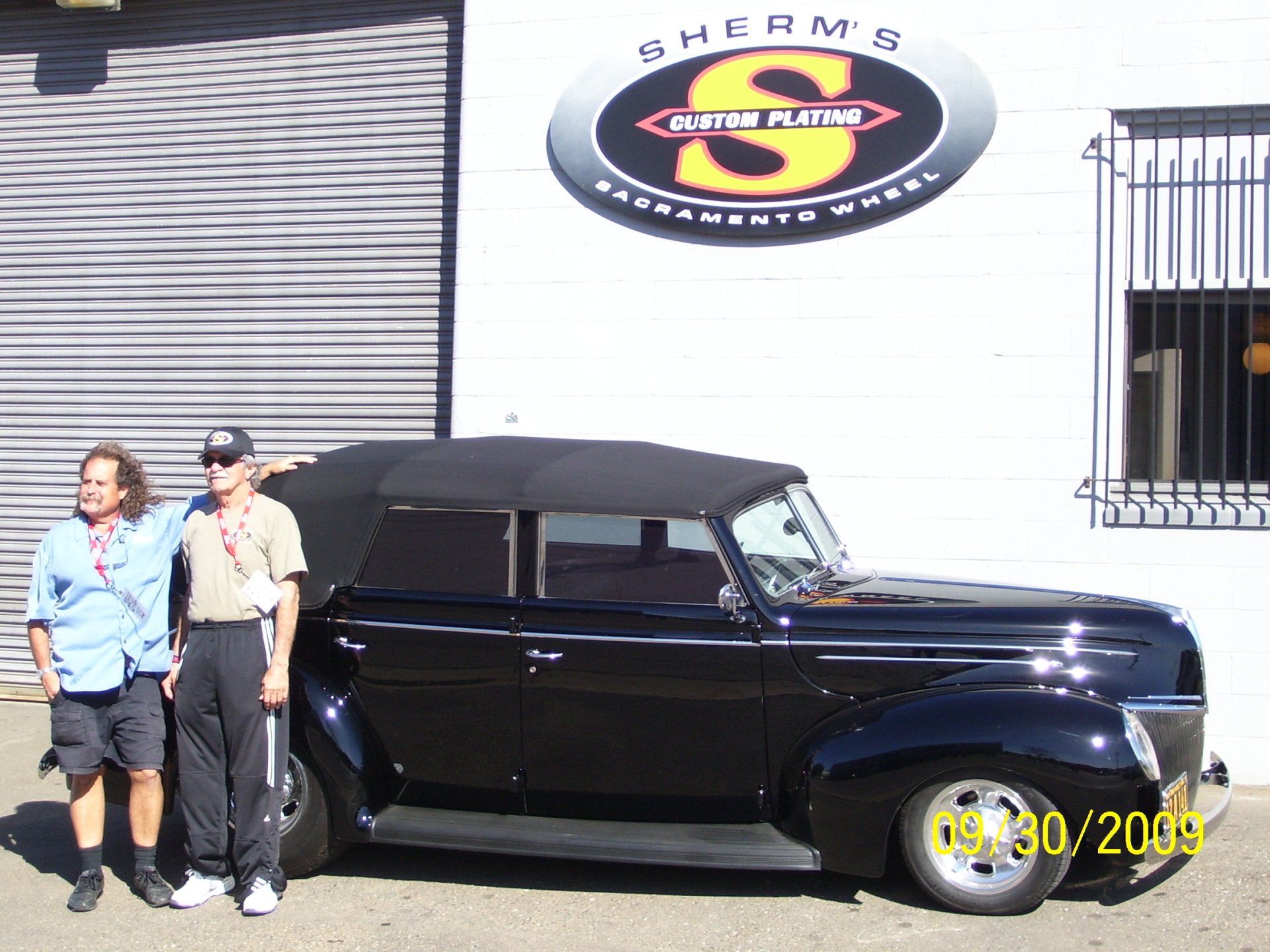 Two men standing next to a black car in front of a sign that says sherm 's custom plating