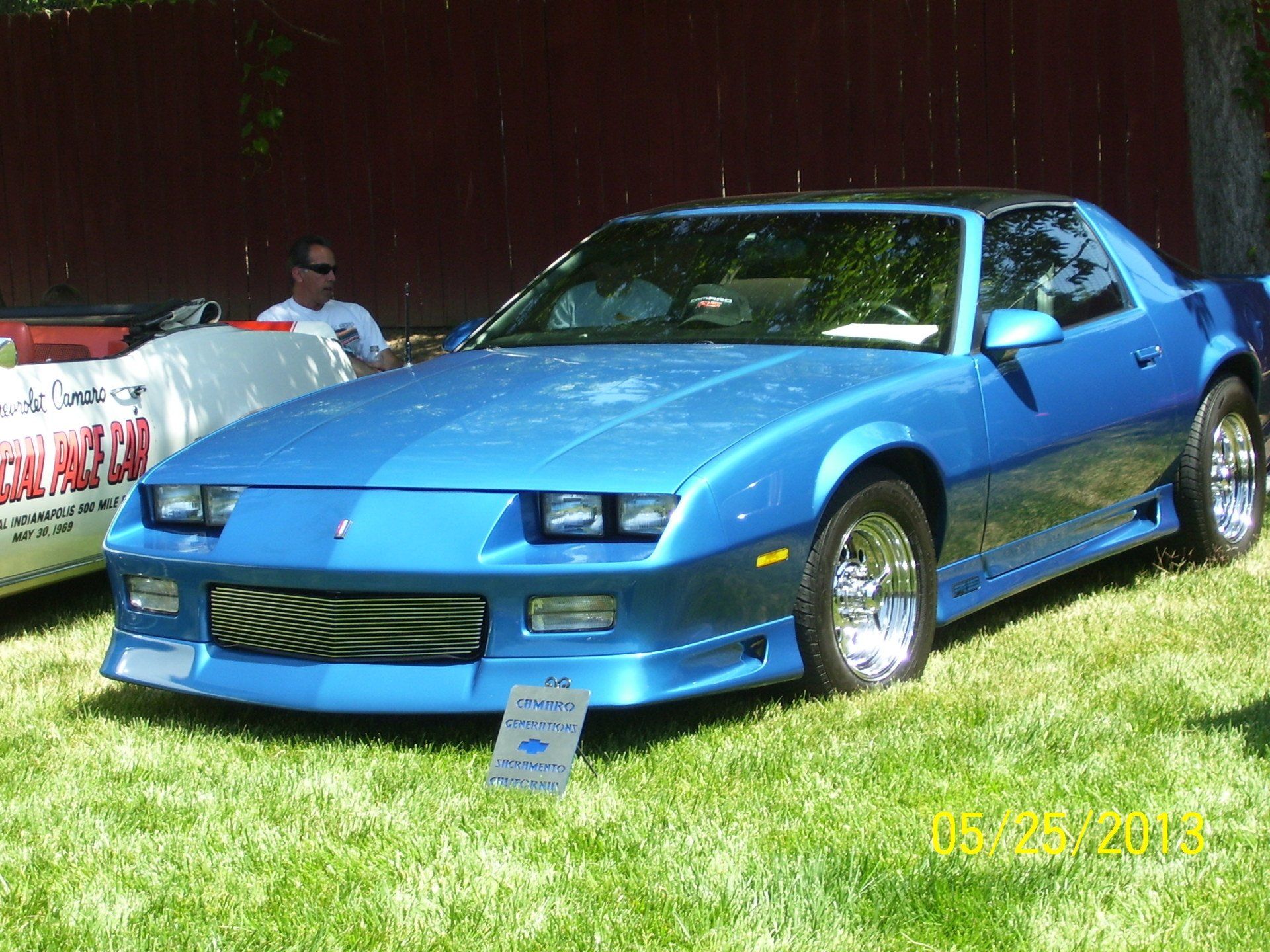 A blue car is parked in the grass with a man sitting in the background