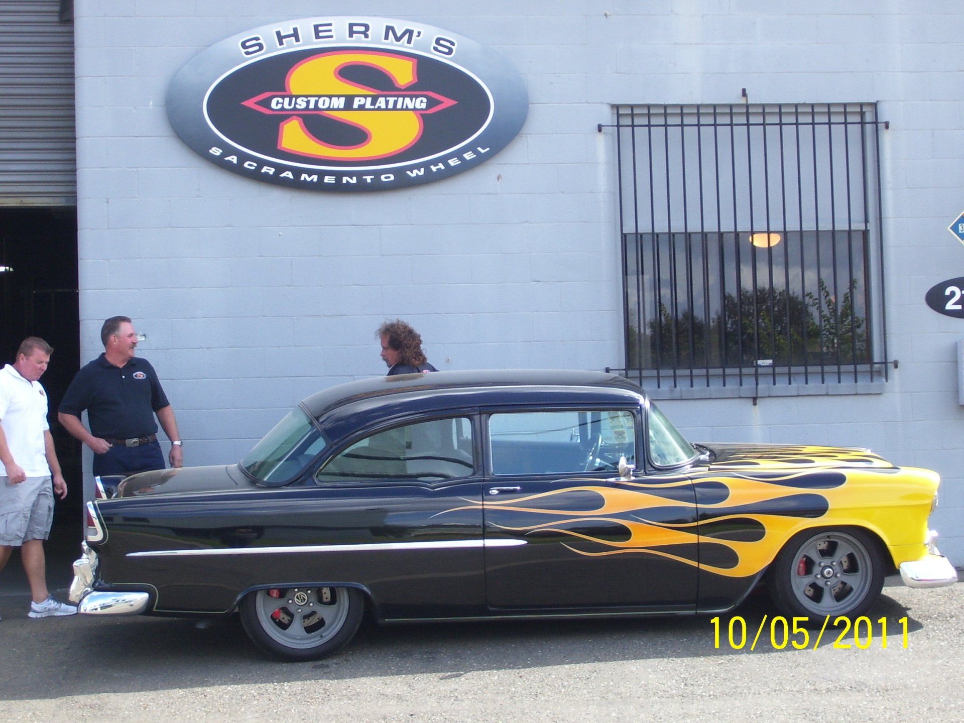 A black and yellow car is parked in front of a building that says sherms custom plating
