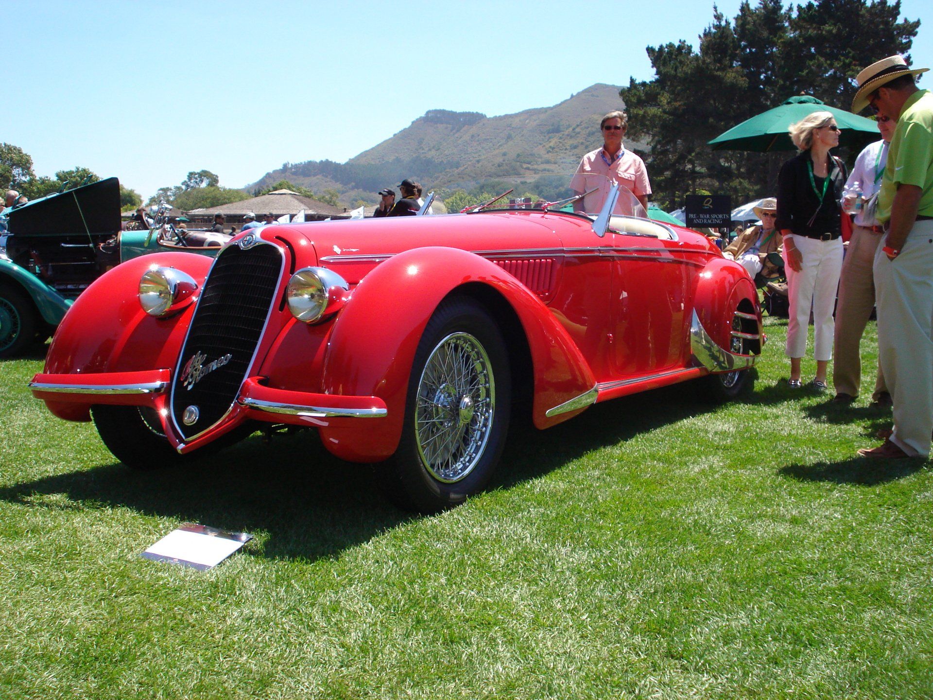 A red car is parked in the grass with people standing around it