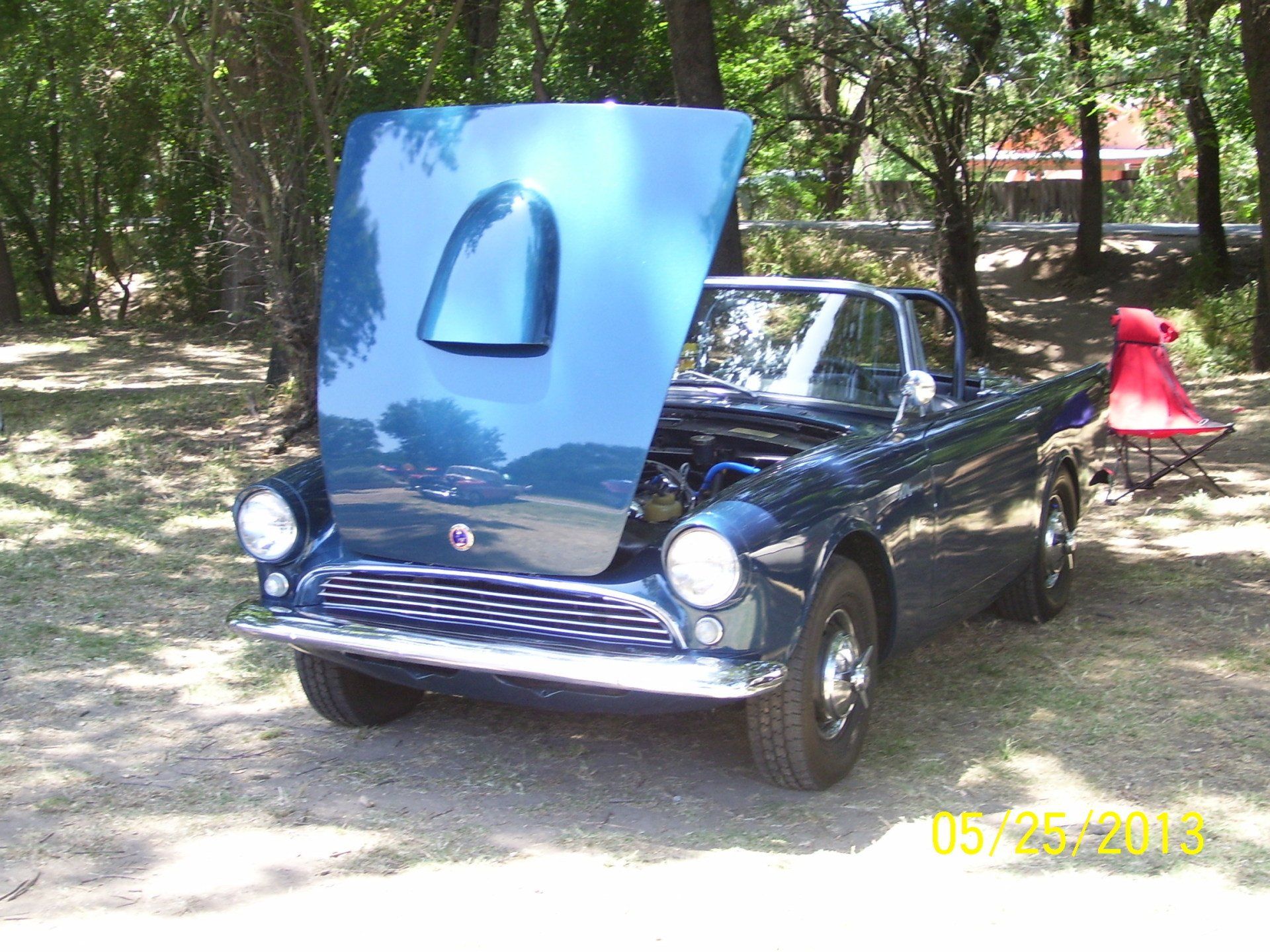 A blue car with the hood up is parked in the woods