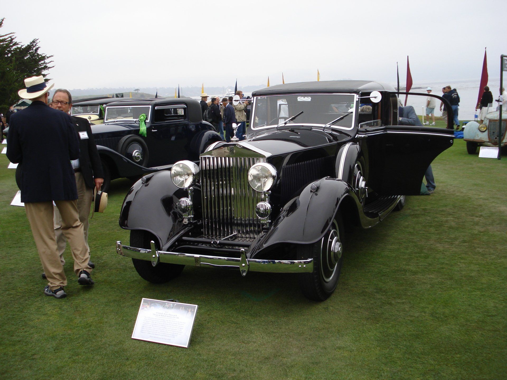A man in a hat stands in front of a black car