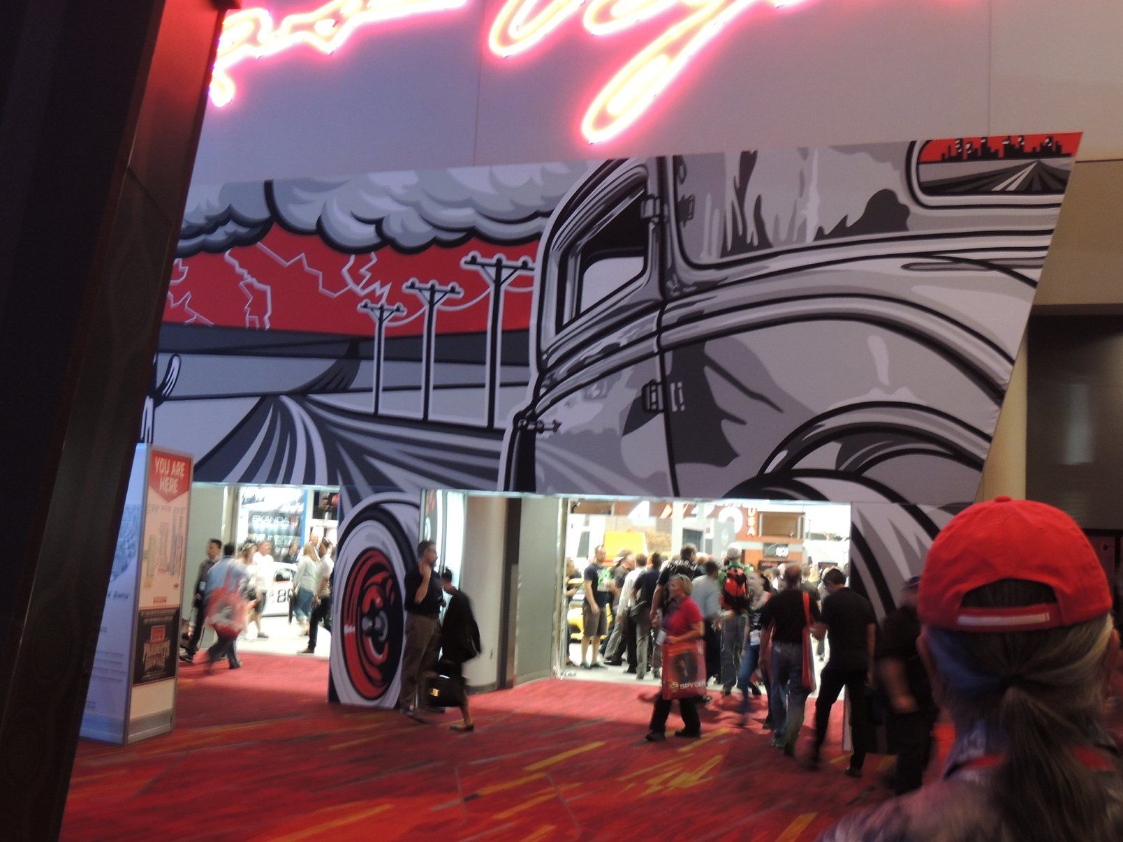 A group of people are walking in a hallway with a large mural on the wall and a neon sign that says 