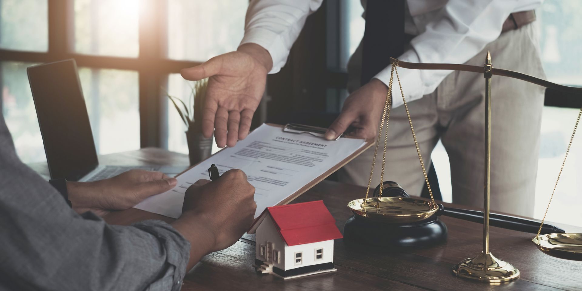 Client signing legal documents with a bankruptcy attorney, scales, and a house model on the desk. Client signing legal documents with a bankruptcy attorney, scales, and a house model on the desk.