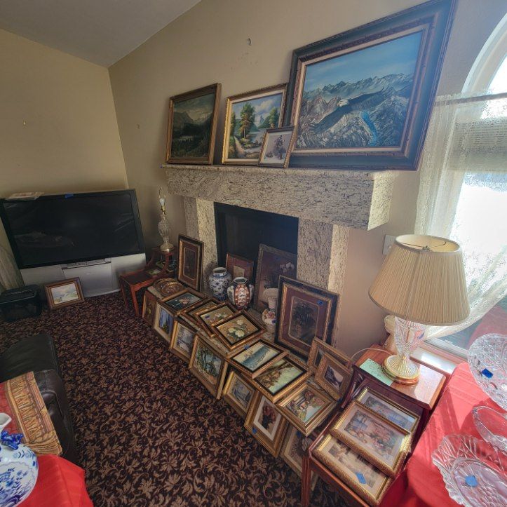 Fireplace mantle adorned with art. Numerous framed pictures line the floor. Lamp sits next to a window.