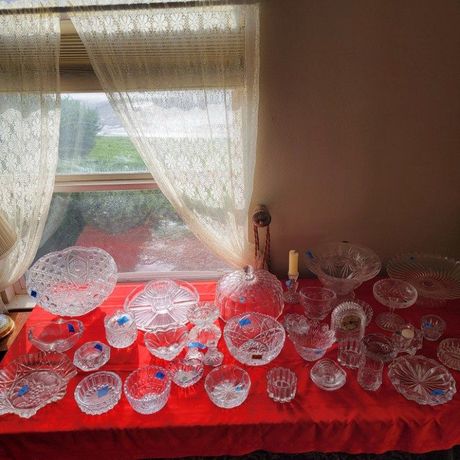 A collection of cut crystal bowls and dishes on a red tablecloth in front of a window with lace curtains.