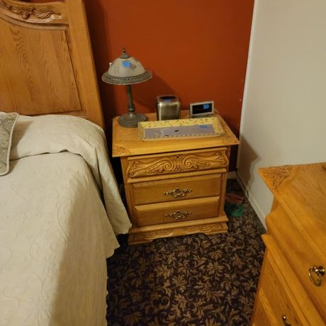 Wooden nightstand beside a bed; lamp, alarm clock, and remote on top.