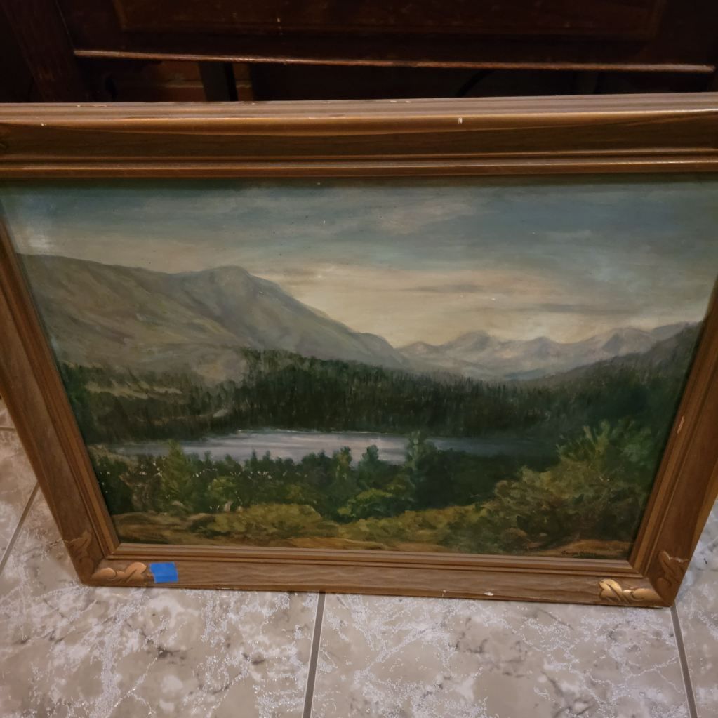 Landscape painting of mountains, a lake, and trees, framed in gold.