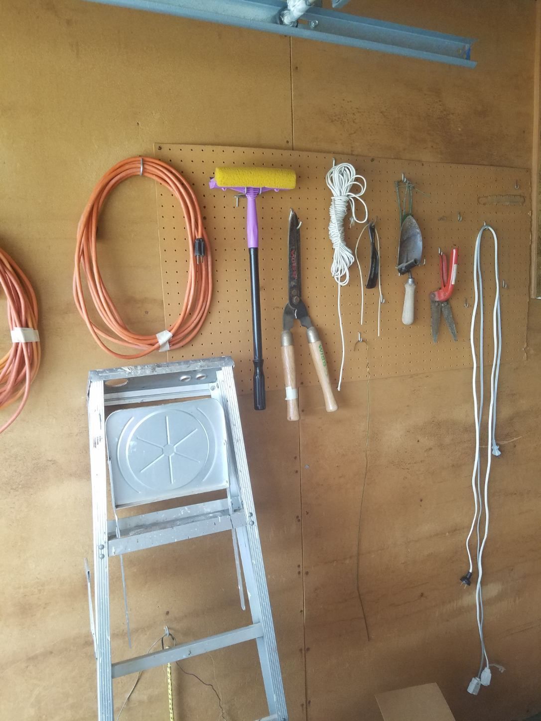 Garage wall with tools and a ladder. Orange extension cords, gardening tools, and a step ladder are visible.
