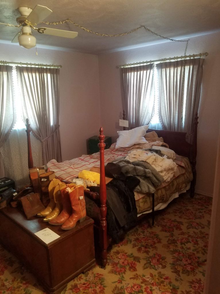 Bedroom with a bed, curtains, chest, boots, and floral carpet.