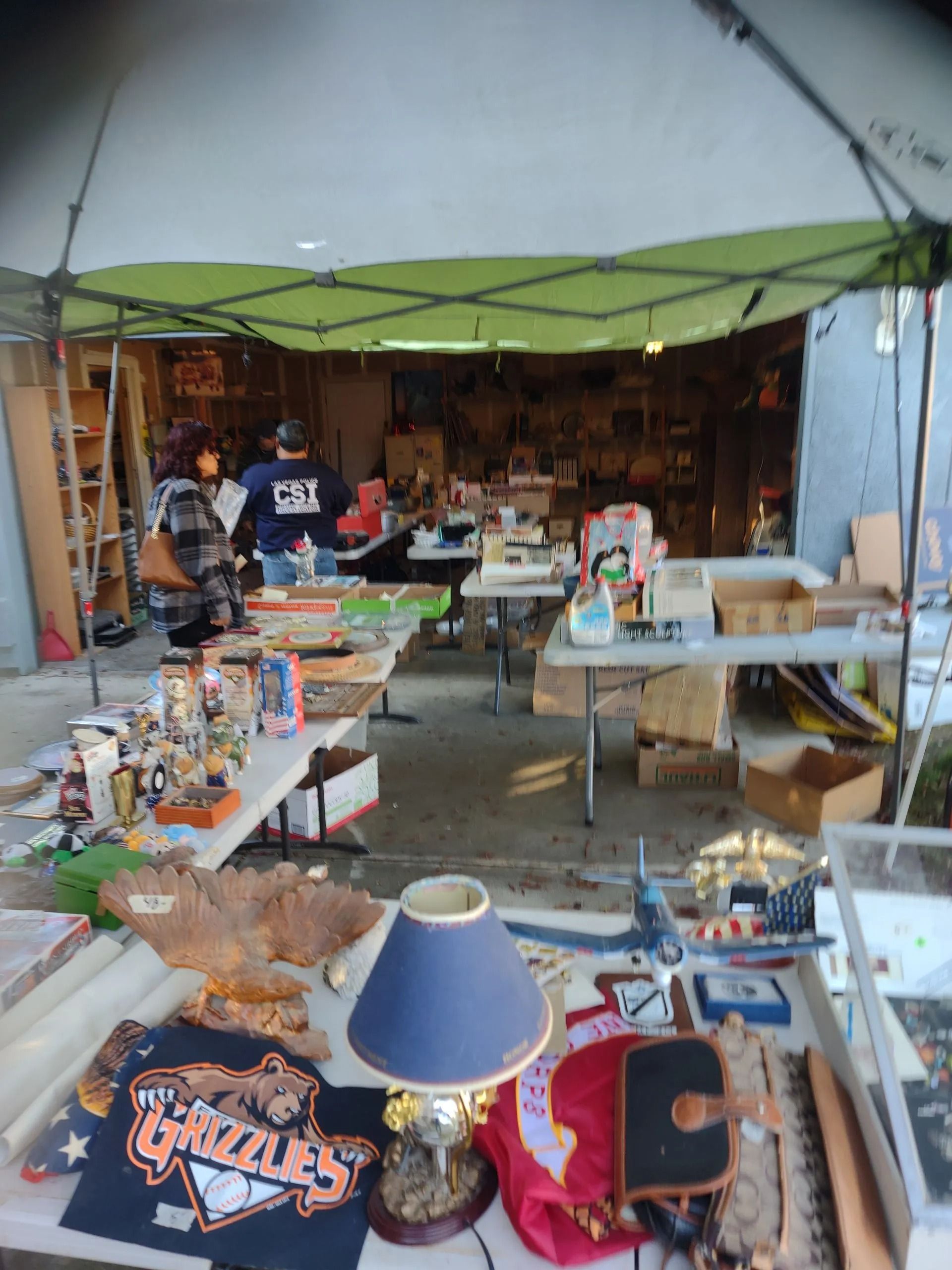 Garage sale scene with tables of items under a canopy. People browse; a man in a Grizzles shirt.