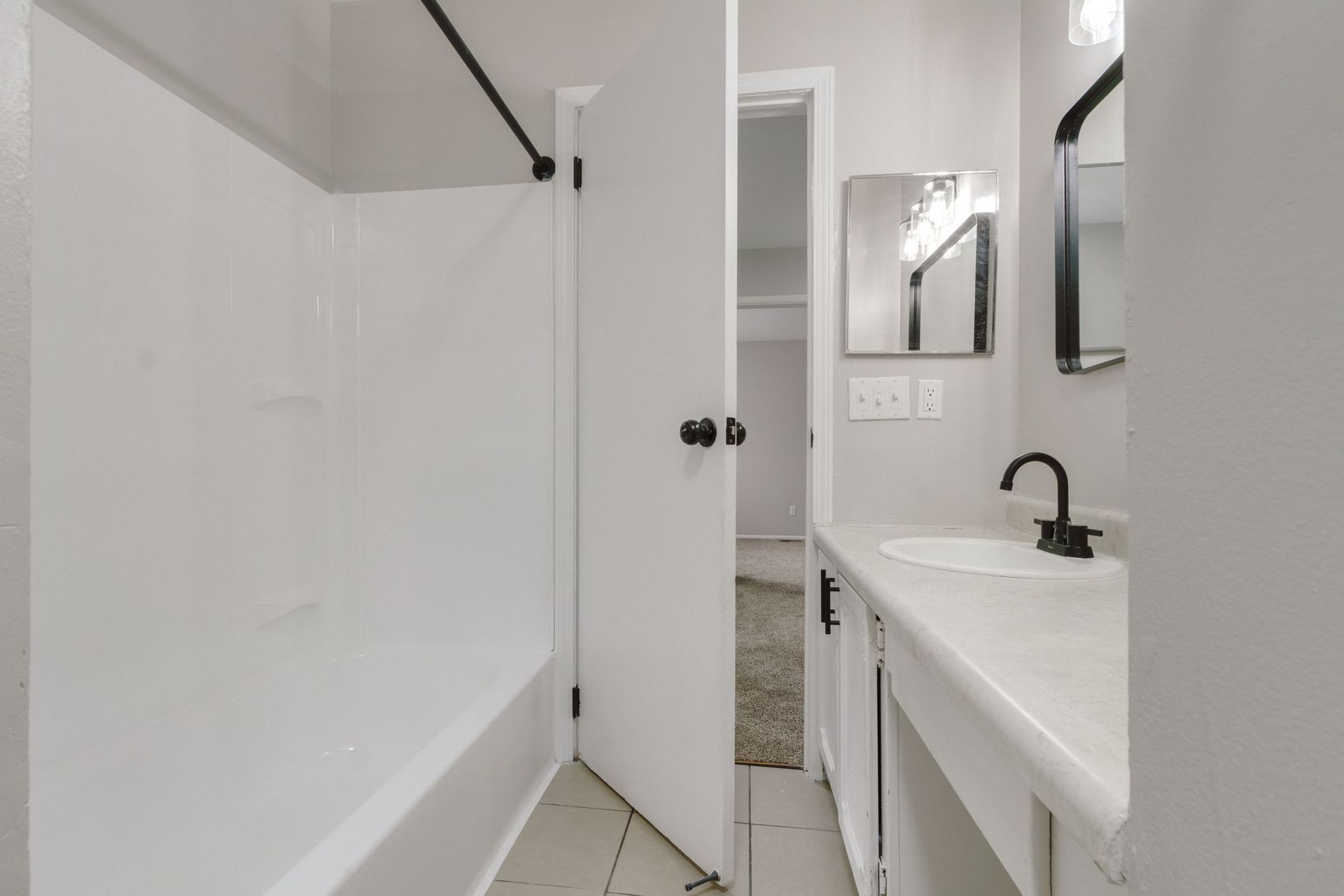 Bathroom with white tub, vanity, and door, black fixtures, and a view into a hallway.
