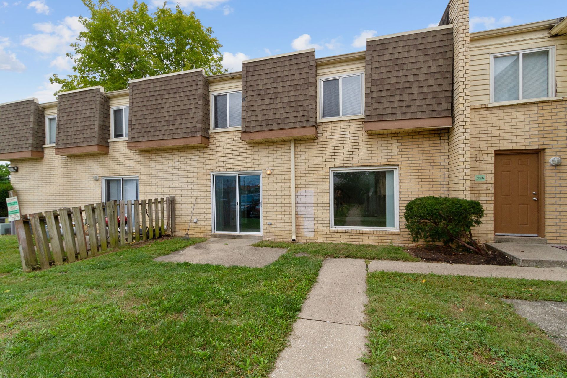 Row of tan townhouses with brown roofs, a green lawn, and a wooden fence.