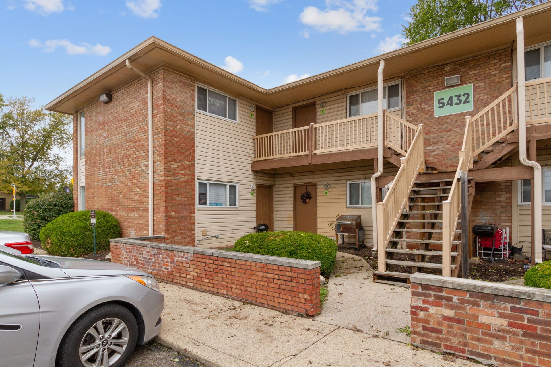 Two-story apartment building with brick and beige siding. Stairs lead to second-floor units. A silver car is parked in front.