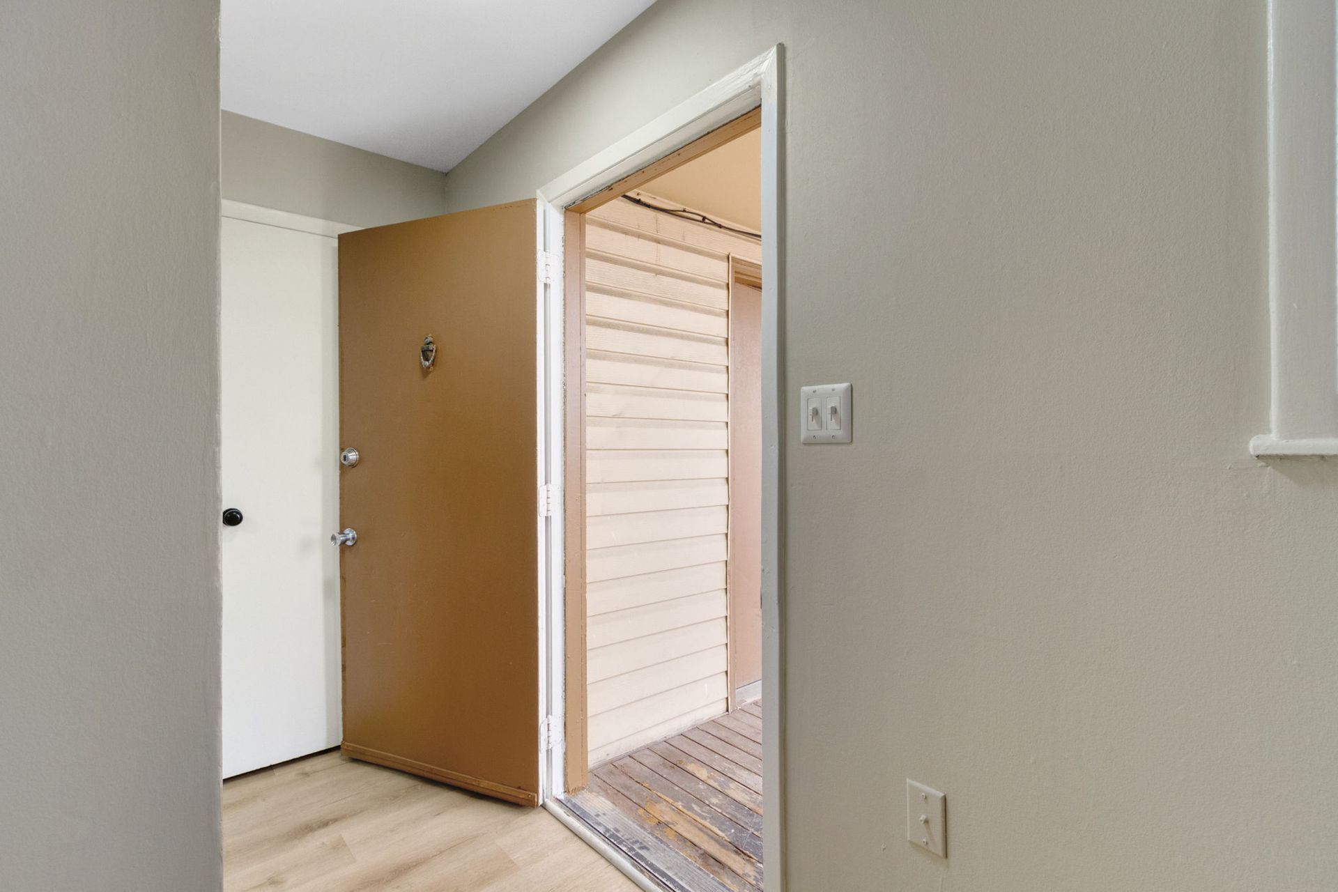 Apartment entryway with open brown door, beige walls, and a view of an outdoor walkway.