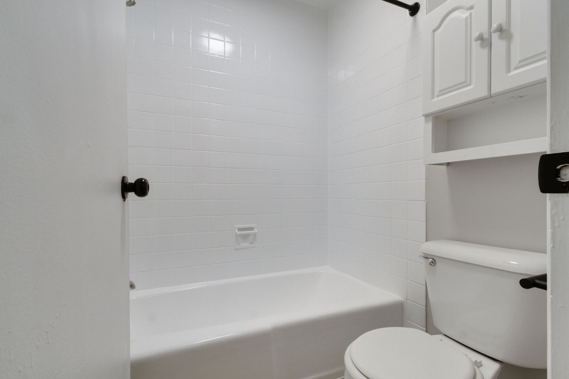White tiled bathroom with tub, toilet, and cabinet. Black door hardware.