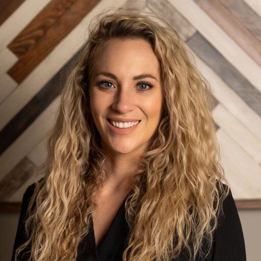 A woman with long blonde hair is smiling in front of a wooden wall.