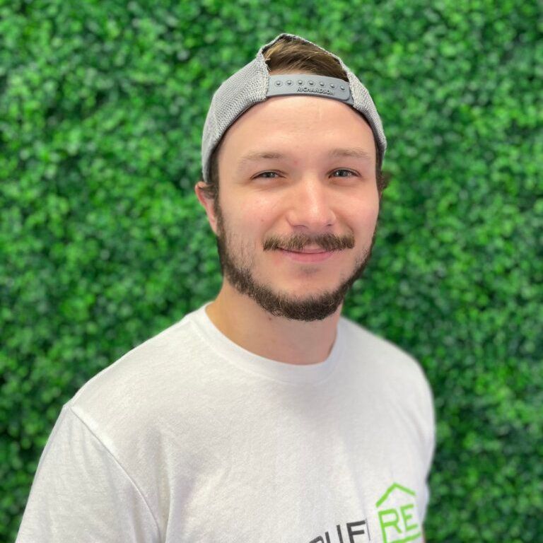 A man wearing a hat and a white shirt is standing in front of a green wall.