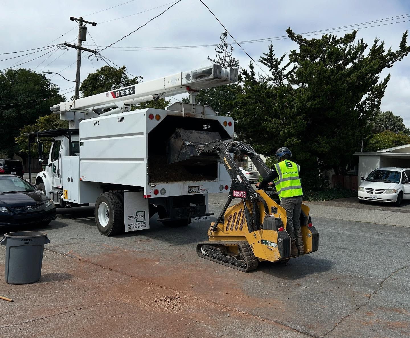 A utility worker in a yellow vest operates a small loader to clear debris from a truck in a residential area.