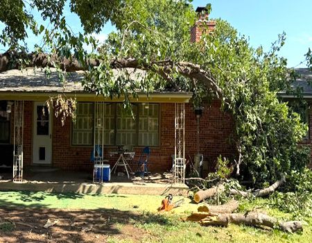 Tree branch fallen on red brick house, chainsaw on ground, sunny day.
