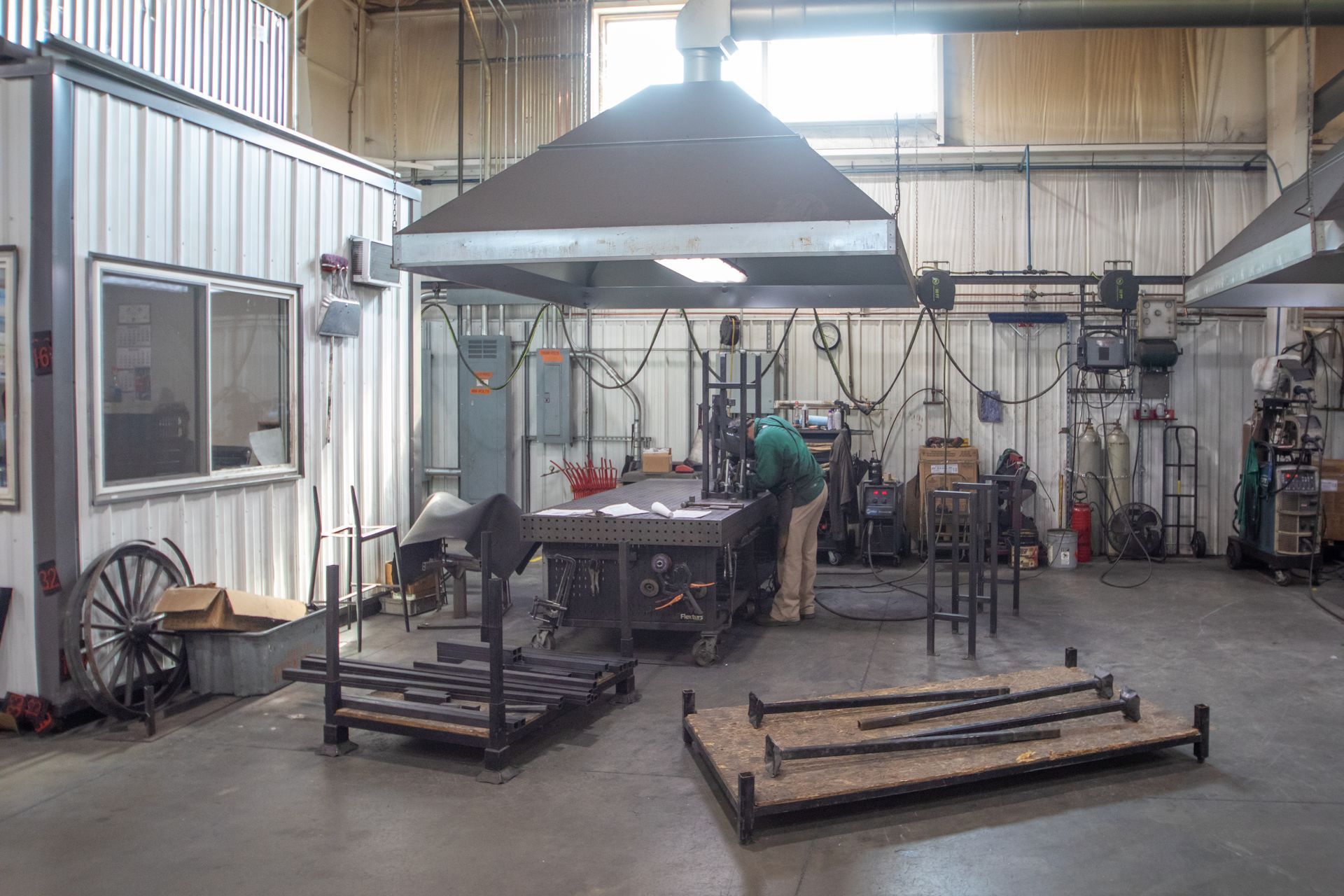 A blacksmith working at a workbench under a fume hood in a workshop, surrounded by tools and metalwork.
