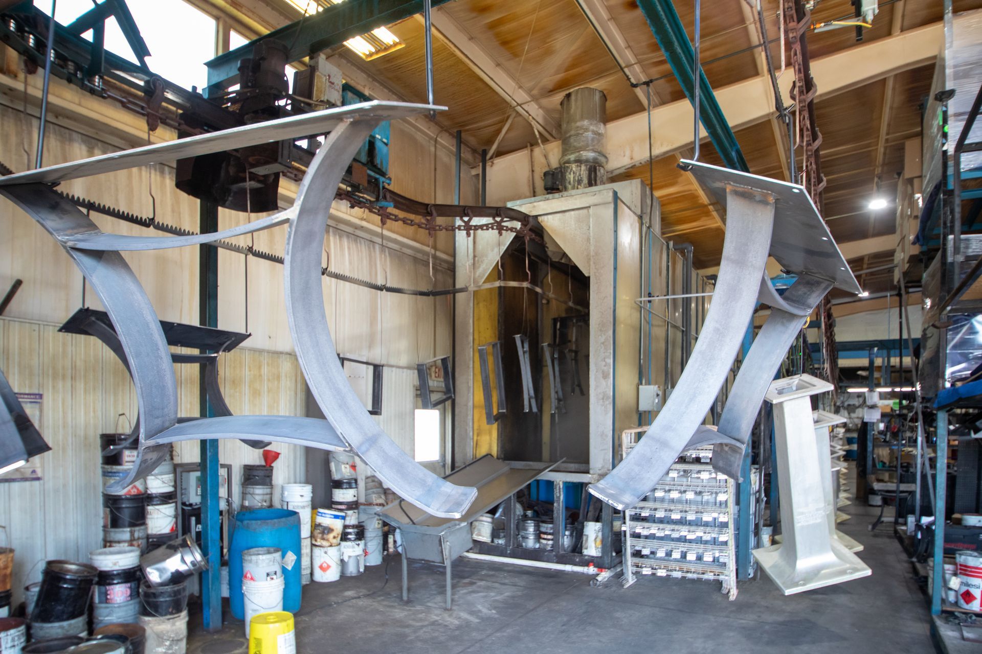 Metal parts hanging in a factory, likely for painting. A large spray booth is in the background.