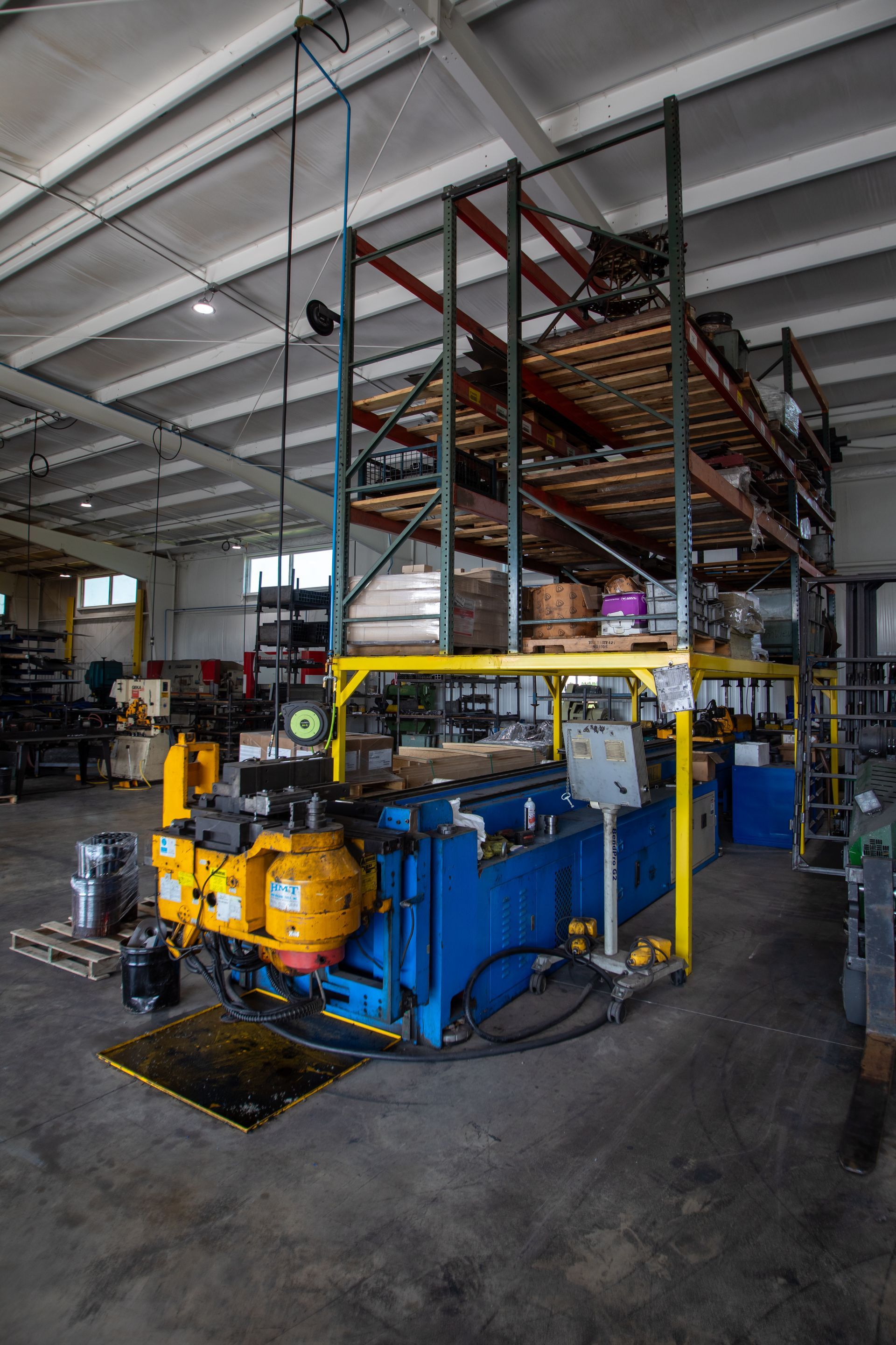 Blue industrial bending machine in a warehouse with shelving unit.