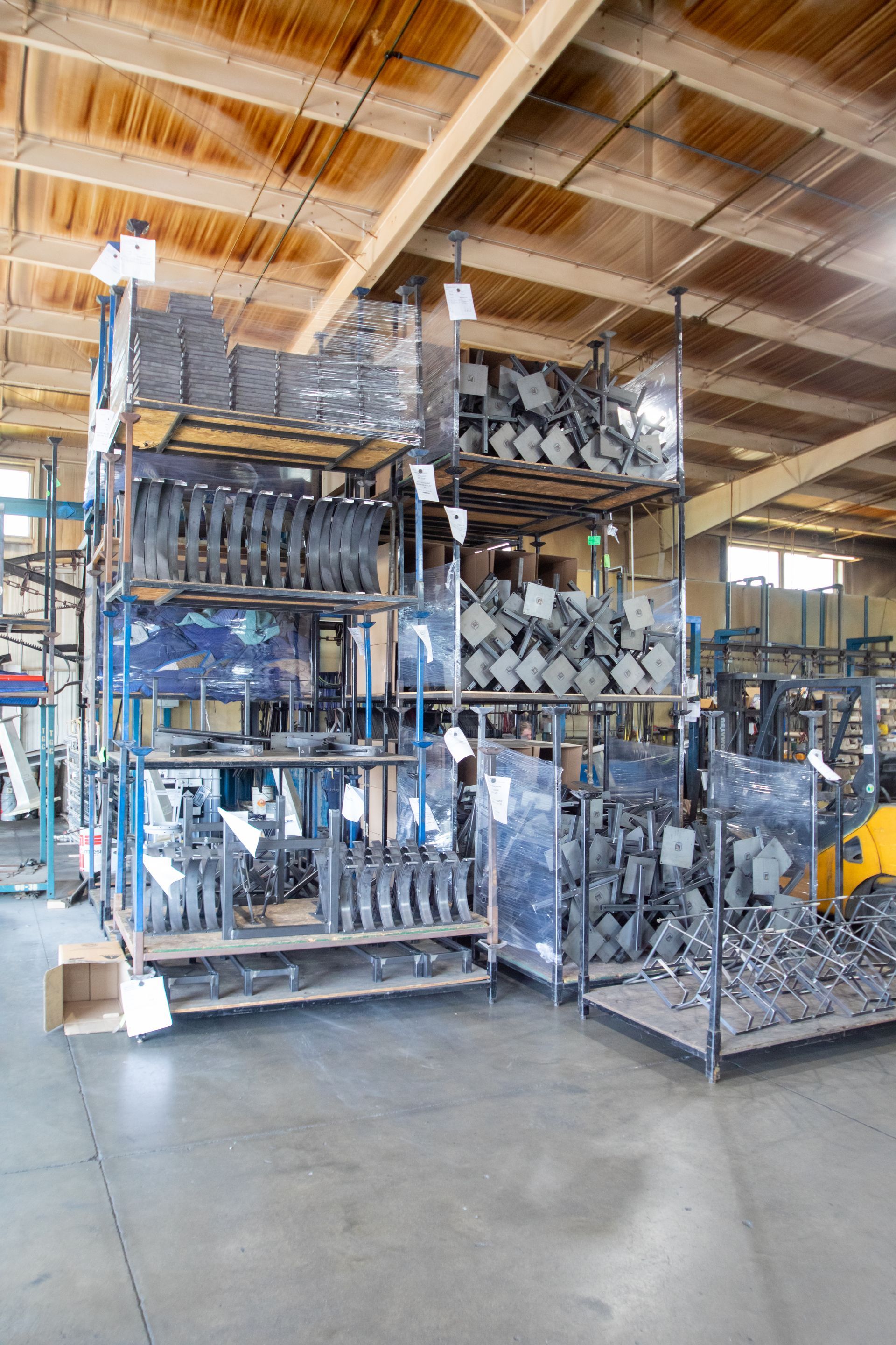 Shelves stacked with gray metal parts in a warehouse. Fluorescent lighting, wooden rafters, and a forklift.