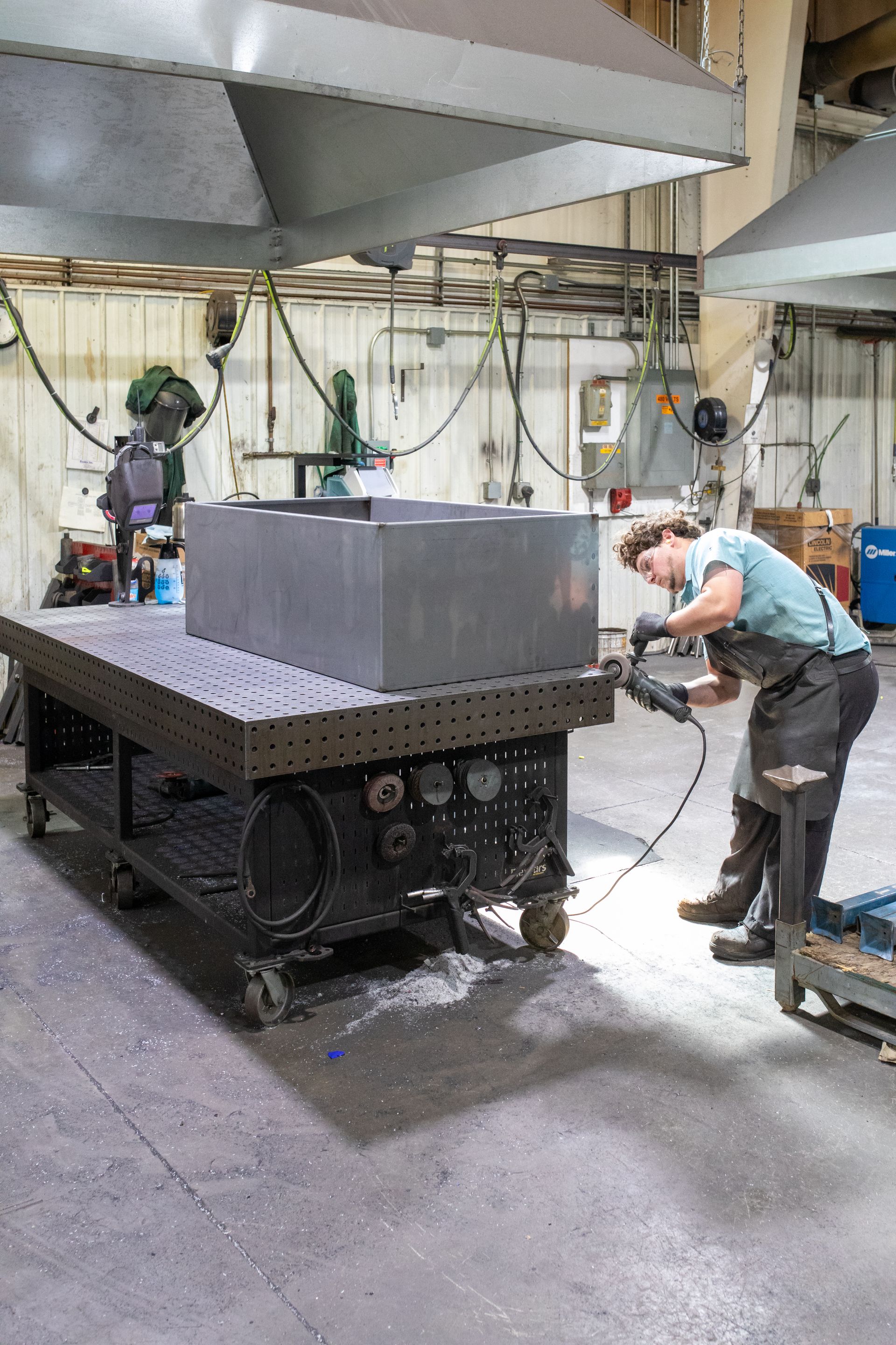 Person grinding metal box on a large, metal workbench in a workshop.