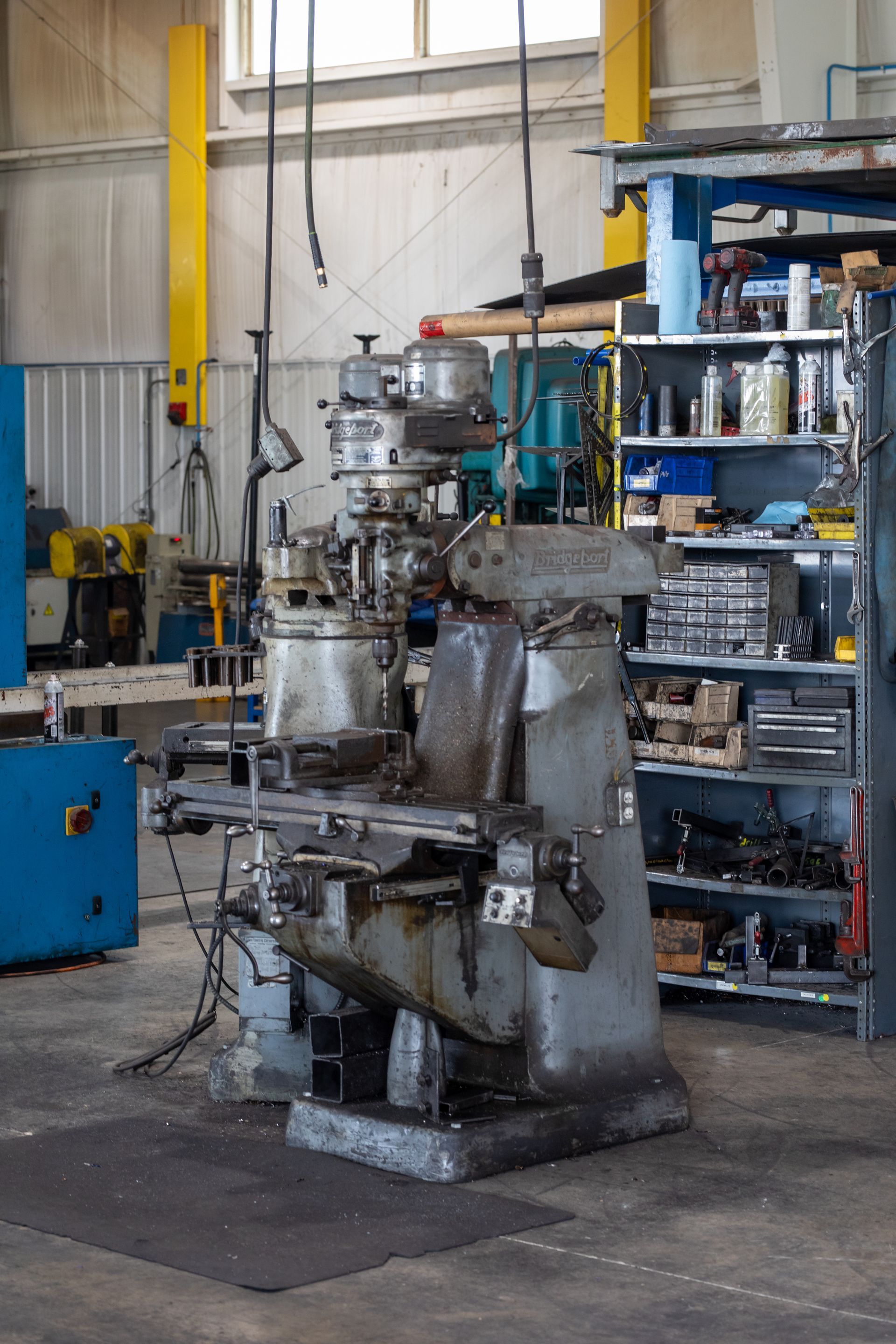 A milling machine in a workshop, grey with some rust. Tools and shelves are in the background.