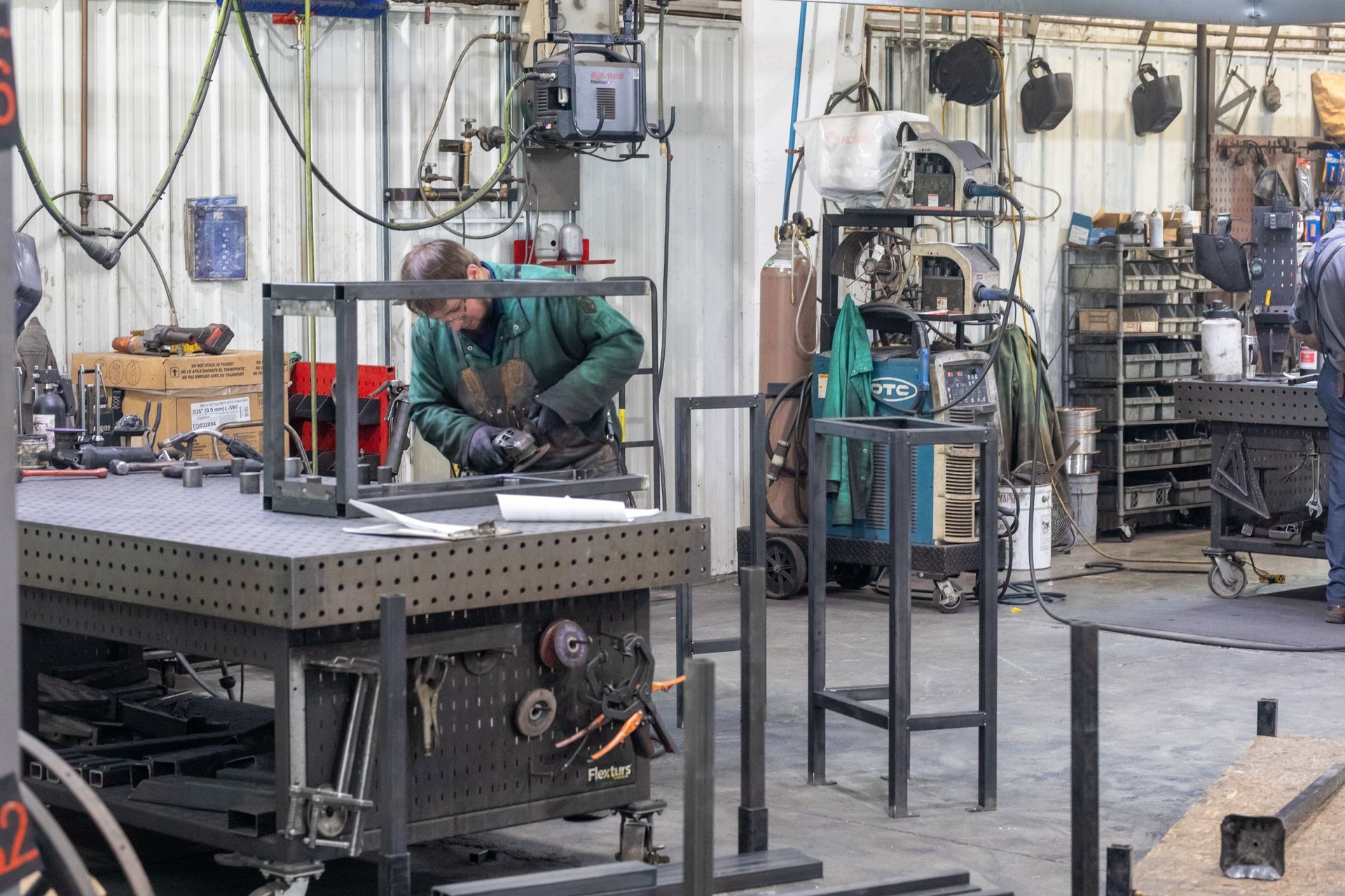 Man welding metal frame in a workshop, with tools and equipment in the background.