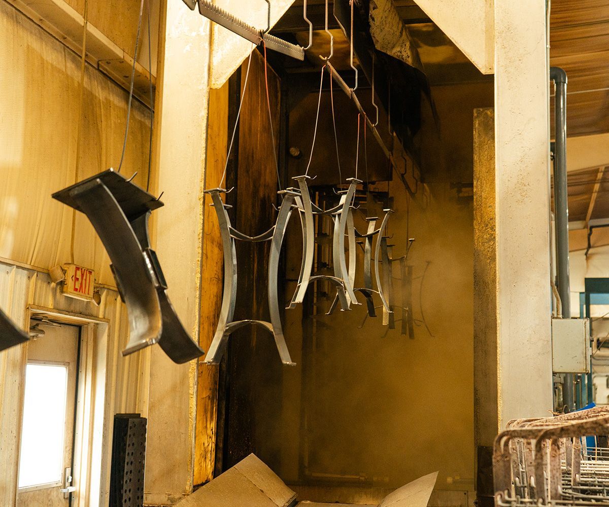 Metal frames hanging on a conveyor in a factory, likely undergoing a finishing process, with visible fumes.