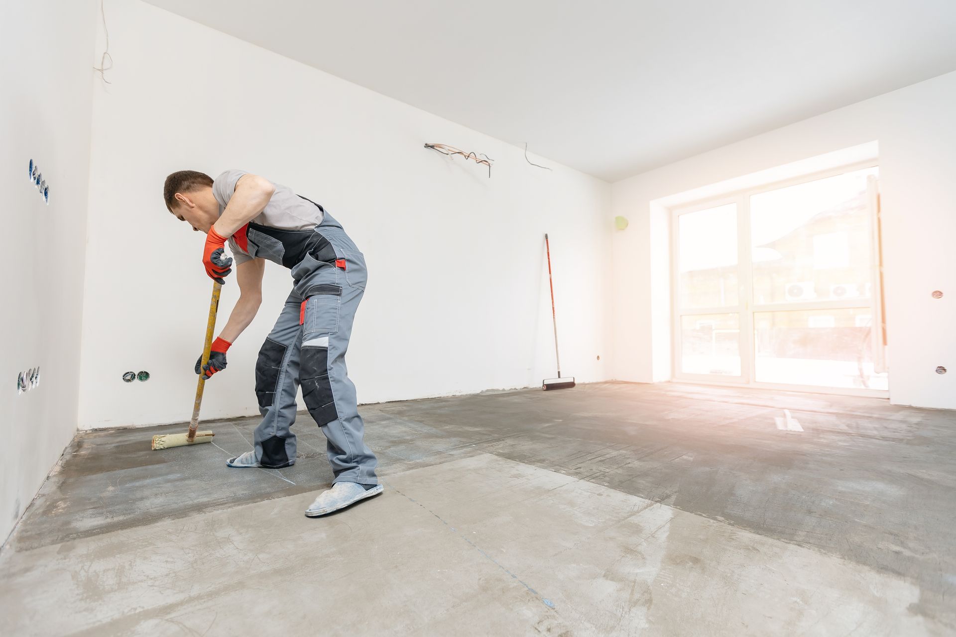 Man in work clothes smoothing cement floor with a long brush in a room under construction.