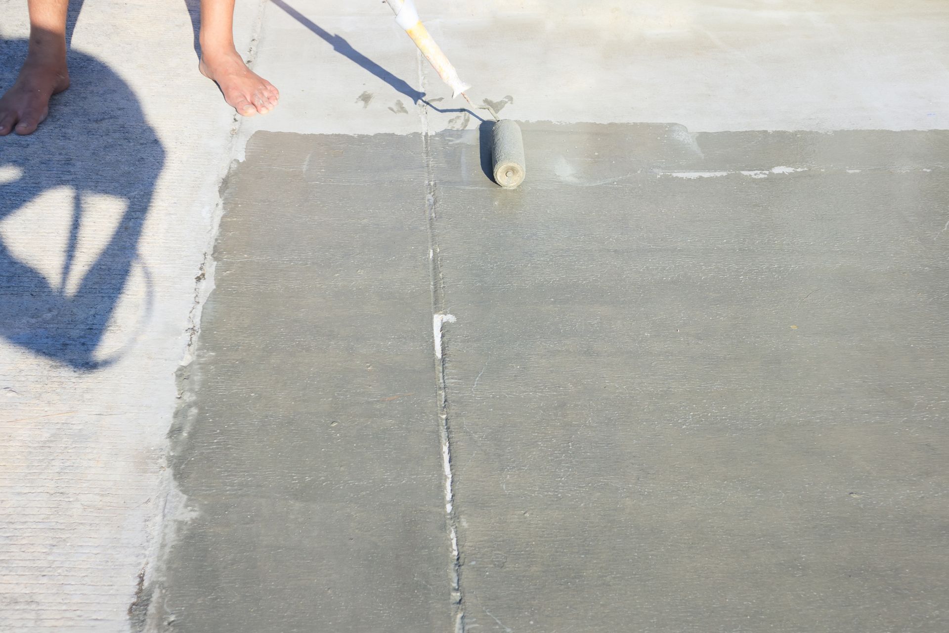 Person applying gray coating to concrete with a roller outdoors. Bare feet in the upper left.