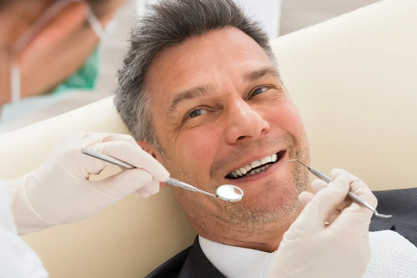 A dentist wearing white gloves uses a mirror and dental tool to examine a patient's teeth in a clinical setting.