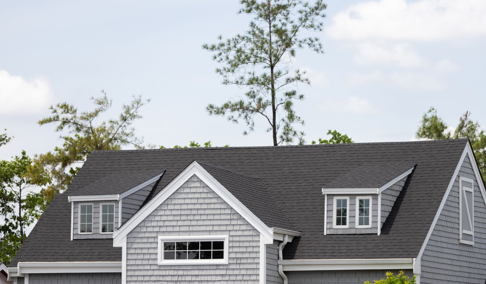 Gray house with shingle roof, dormer windows, and a tree in the background.