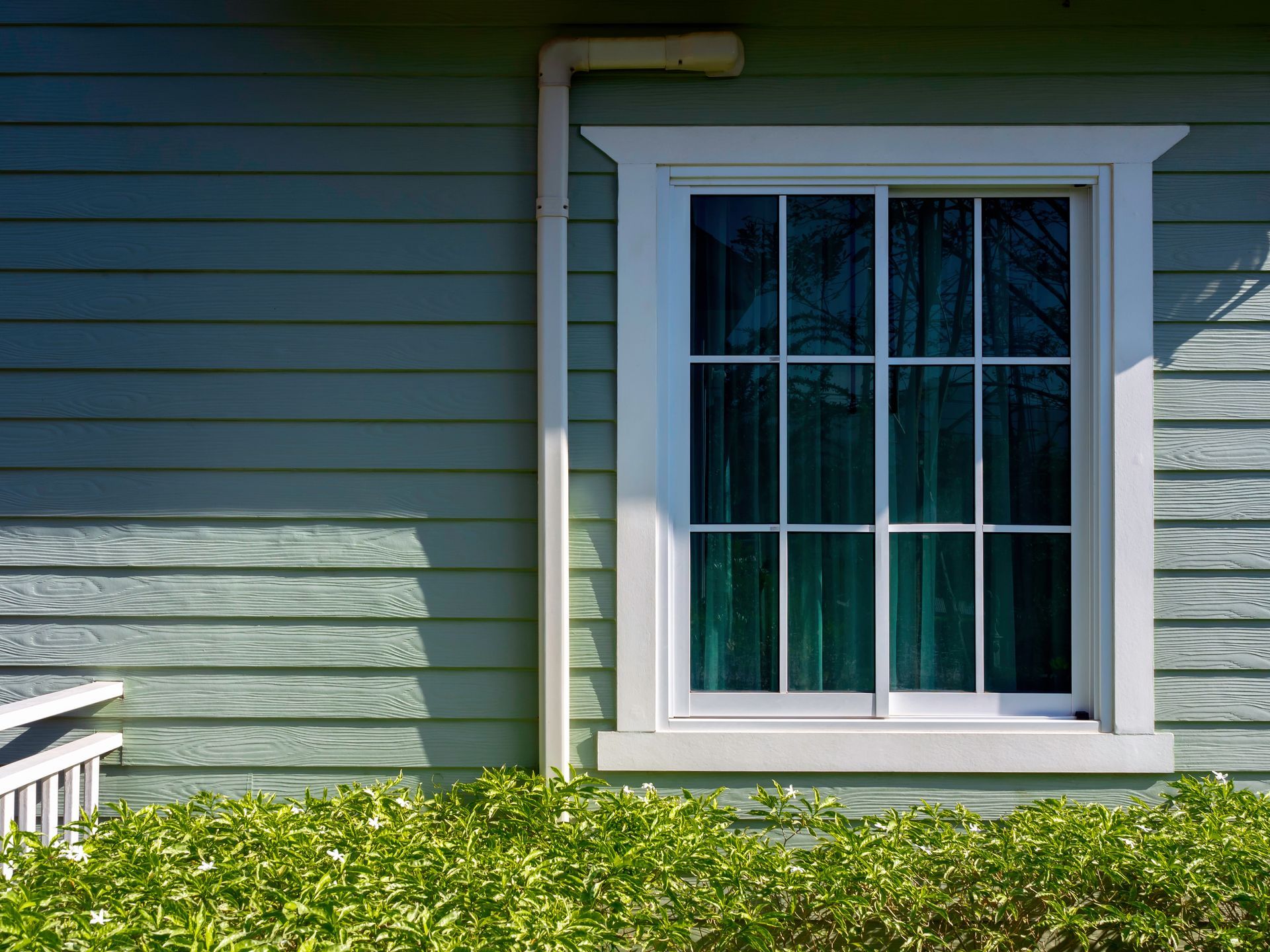 Window on a blue-green house with white trim, green curtains, and a pipe. Green bush at bottom.