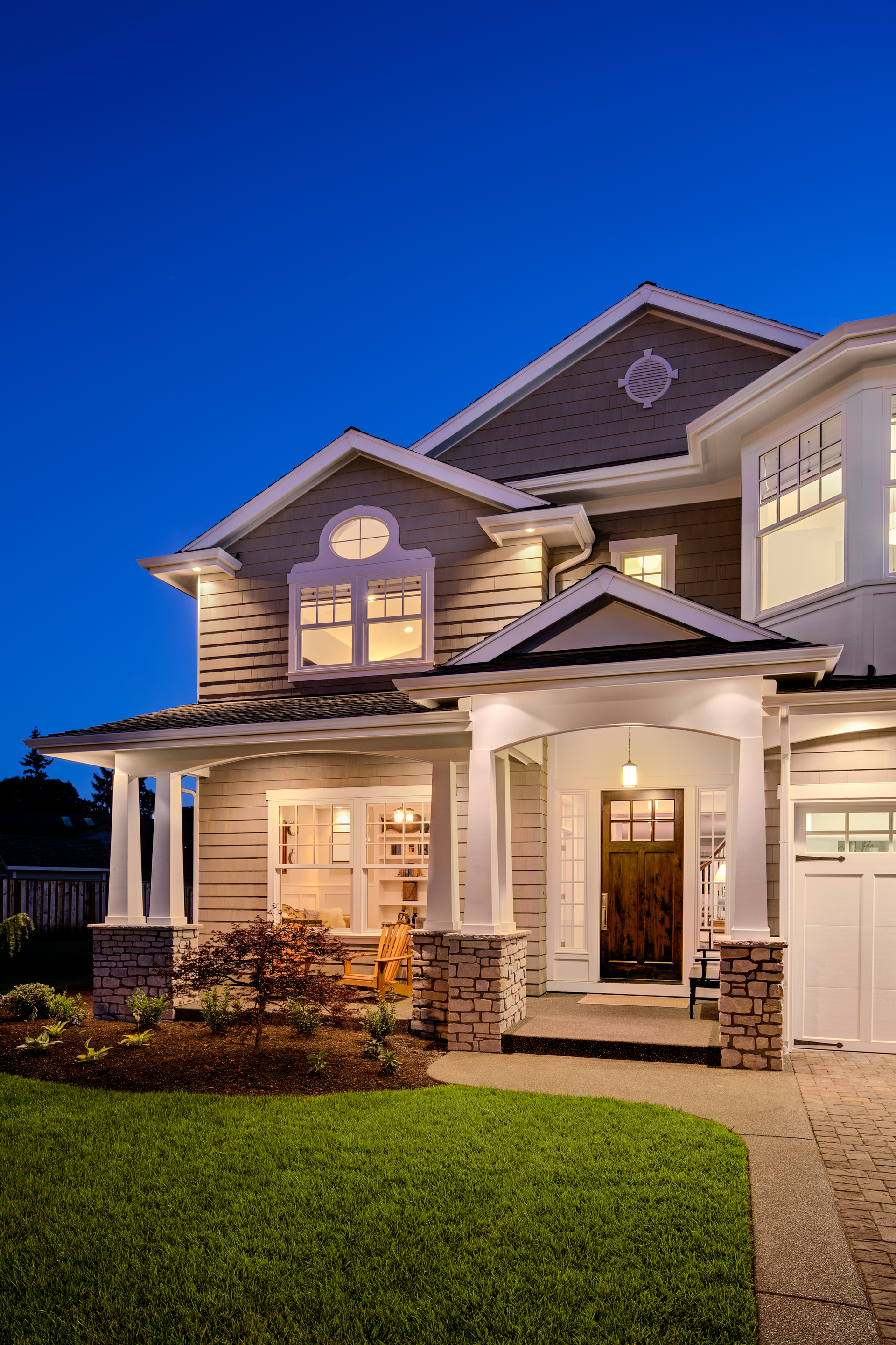 Two-story house with illuminated windows and front porch at dusk; gray siding, stone accents, green lawn.