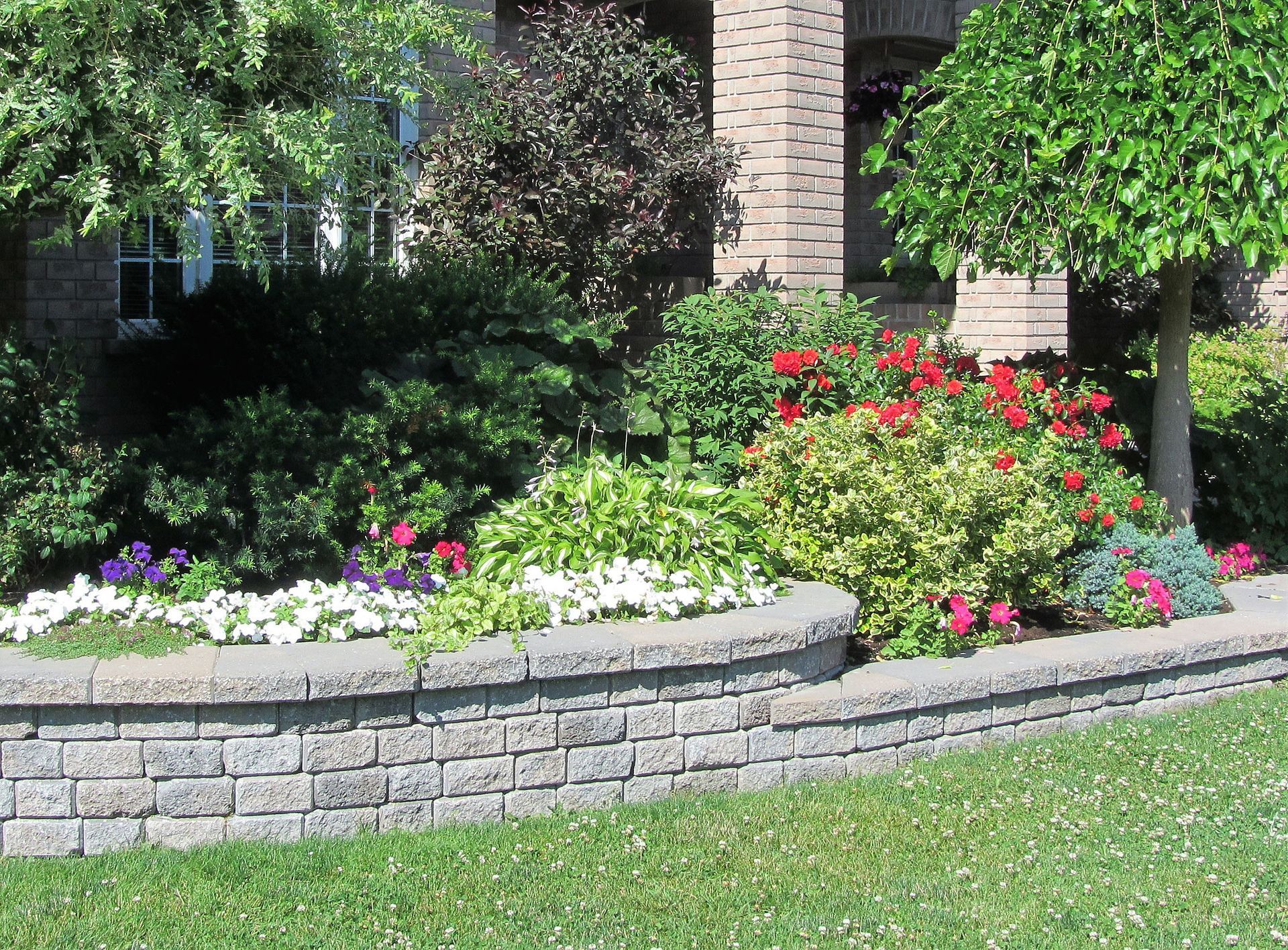 Stone retaining wall with colorful flowers and shrubs in front of a brick house. Stone retaining wall with colorful flowers and shrubs in front of a brick house.