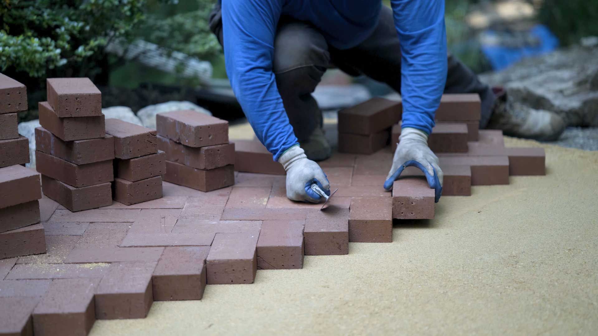 Person laying red bricks in a herringbone pattern on a sand base, outdoors. Person laying red bricks in a herringbone pattern on a sand base, outdoors.