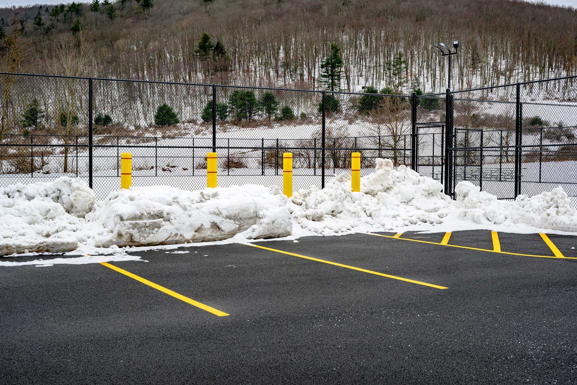 Snow-covered parking lot with yellow painted lines and bollards; a chain-link fence and a snowy forest in the background.