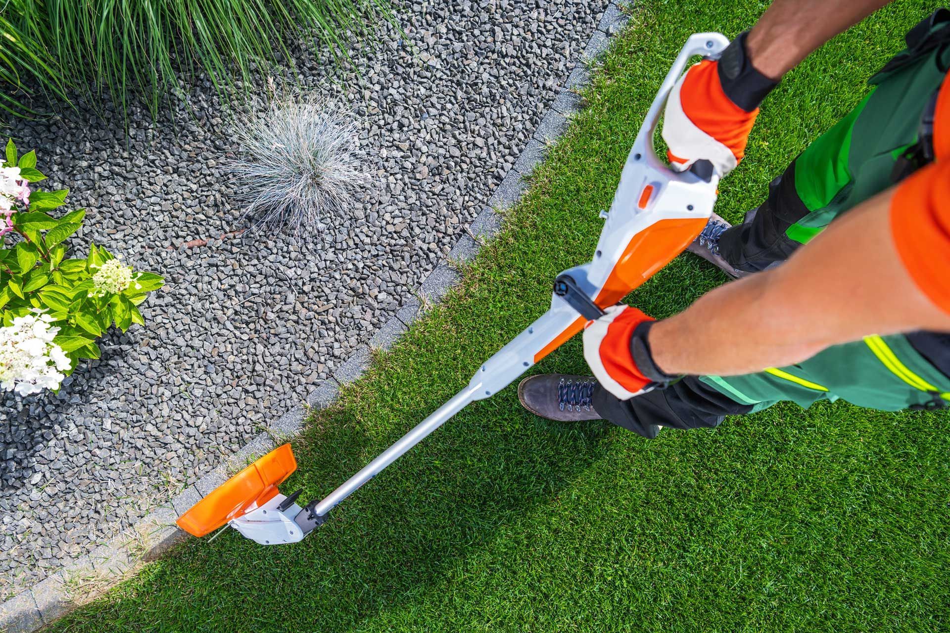 Person edging lawn with an orange and white trimmer next to gravel and flowers. Person edging lawn with an orange and white trimmer next to gravel and flowers.