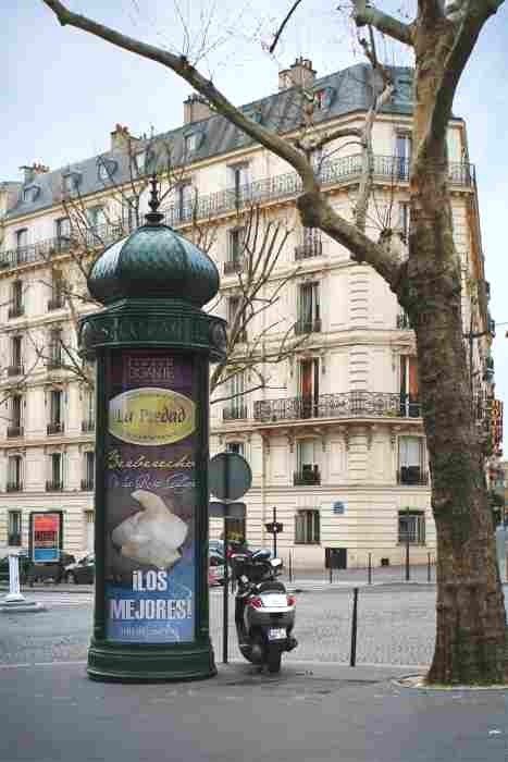 Una columna Morris de color verde oscuro, adornada con carteles publicitarios, se alza en la acera frente a un clásico edificio de apartamentos parisino.