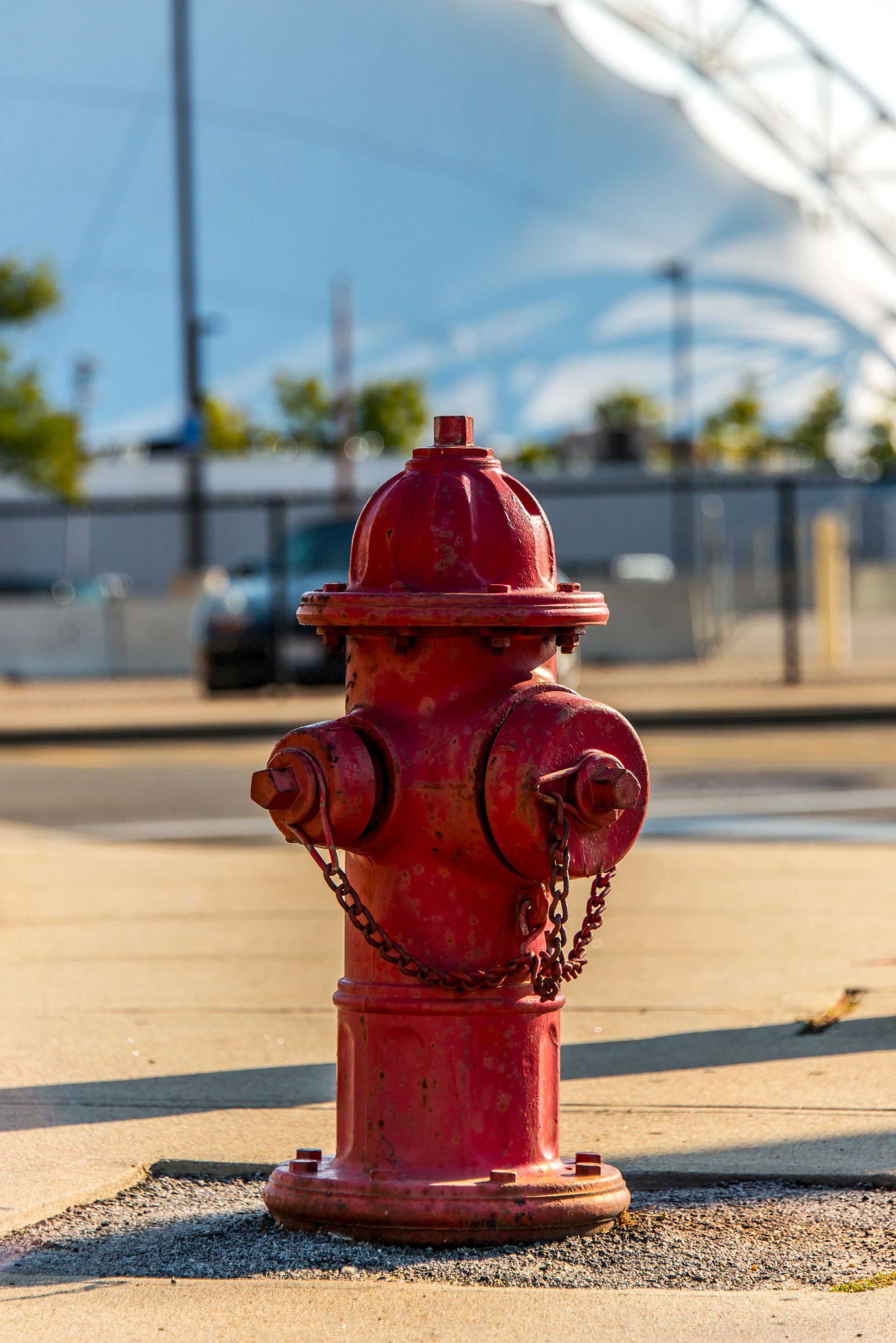Red fire hydrant on concrete, with background including a car, fence, and a white, curved structure.