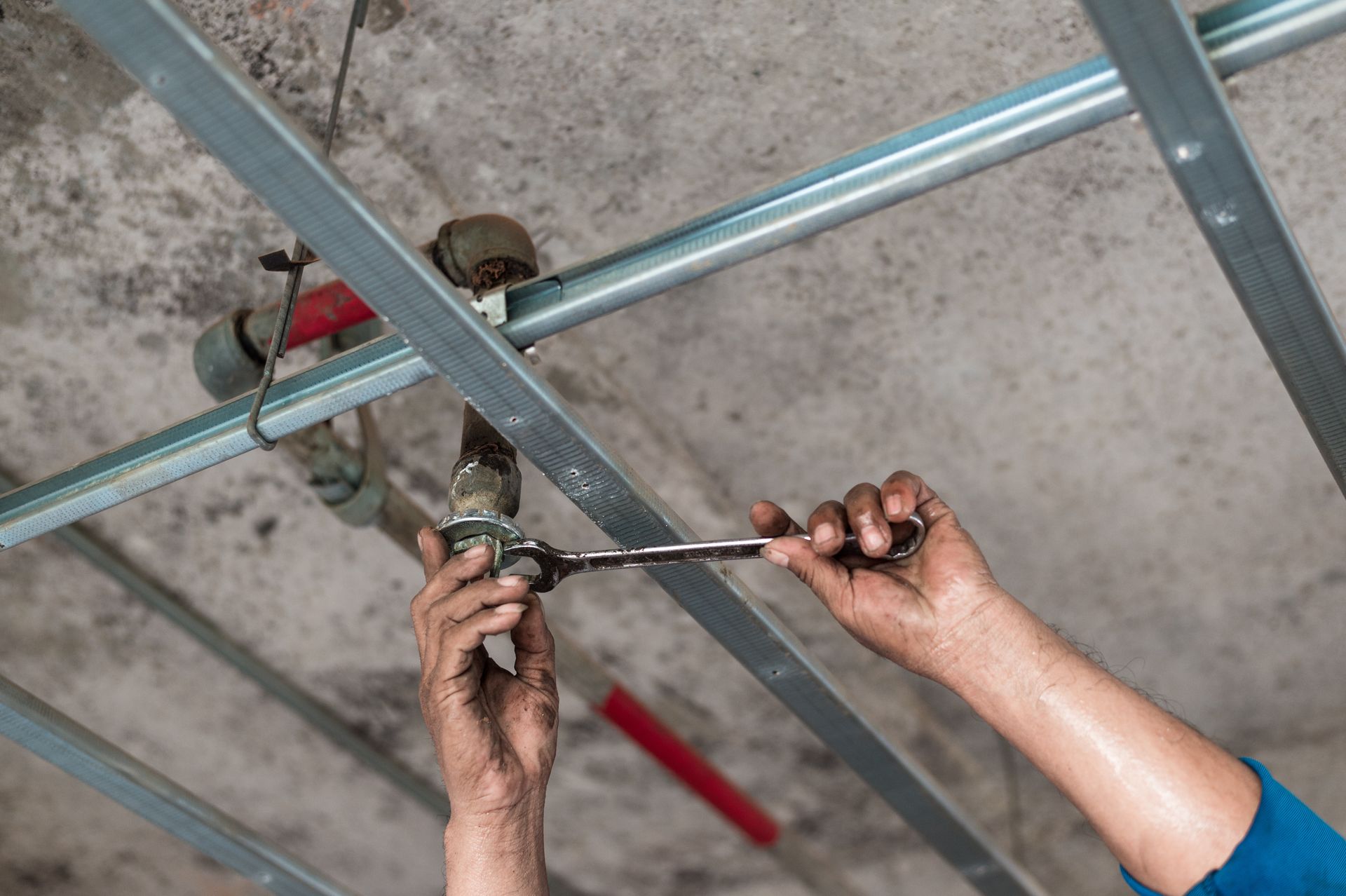 Person using a wrench to work on pipes in a ceiling. Steel beams and concrete ceiling visible.