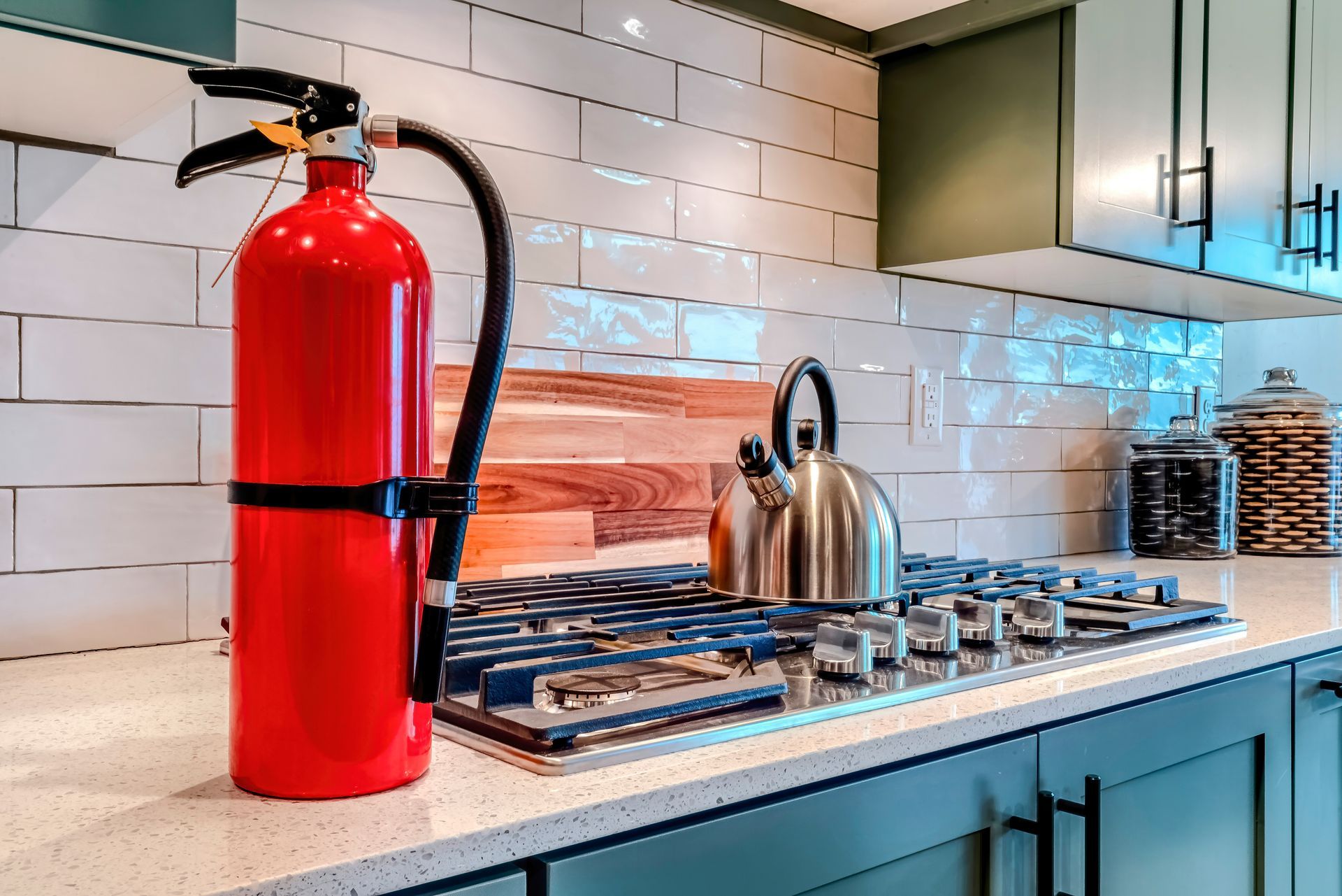 Red fire extinguisher next to a stove in a kitchen with a stainless steel kettle on top.