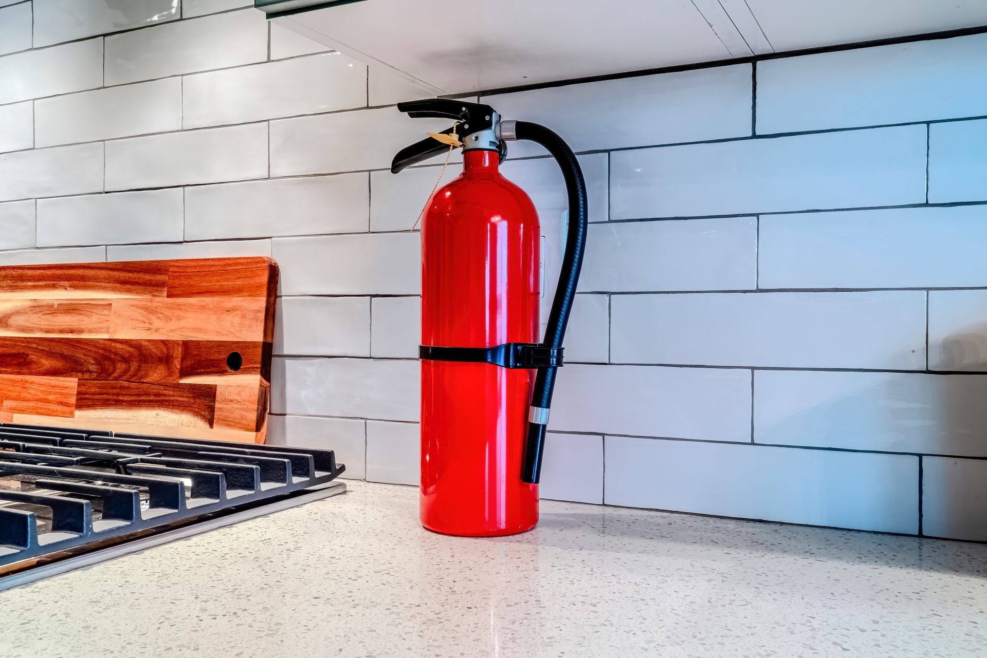 Red fire extinguisher on countertop in a kitchen with white tile backsplash and wooden cutting board. Red fire extinguisher on countertop in a kitchen with white tile backsplash and wooden cutting board.